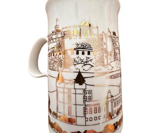May include: White ceramic mug with a gold-colored illustration of a castle and surrounding landscape. The illustration is detailed and includes trees, buildings, and a river.