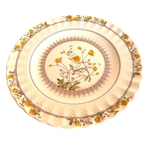 Könnte beinhalten: Ein cremefarbener Keramikteller mit gewelltem Rand. Der Teller hat ein Blumendesign mit gelben Blumen und grünen Blättern in der Mitte, umgeben von einem braunen und cremefarbenen Rand. Der Rand ist mit gelben floralen Akzenten verziert.