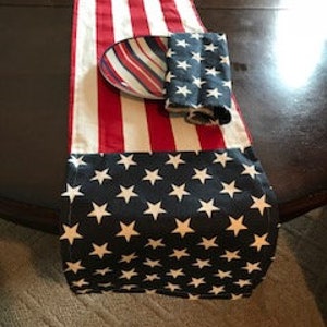 May include: Red, white, and blue table runner with a star pattern on the end. The runner is made of fabric and has a casual, patriotic theme.