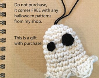 May include: A small, off-white crocheted ghost ornament with two black circular eyes and a black yarn hanging loop, resting on a tan spiral-bound notebook featuring the text "halloween ghost" and additional printed information.