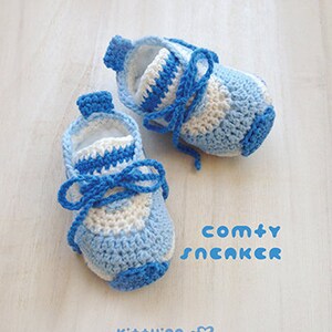 May include: A pair of blue and white crocheted baby shoes with laces. The shoes are on a white wooden surface. The text "comty SNCAKCR" is written in blue letters below the shoes.