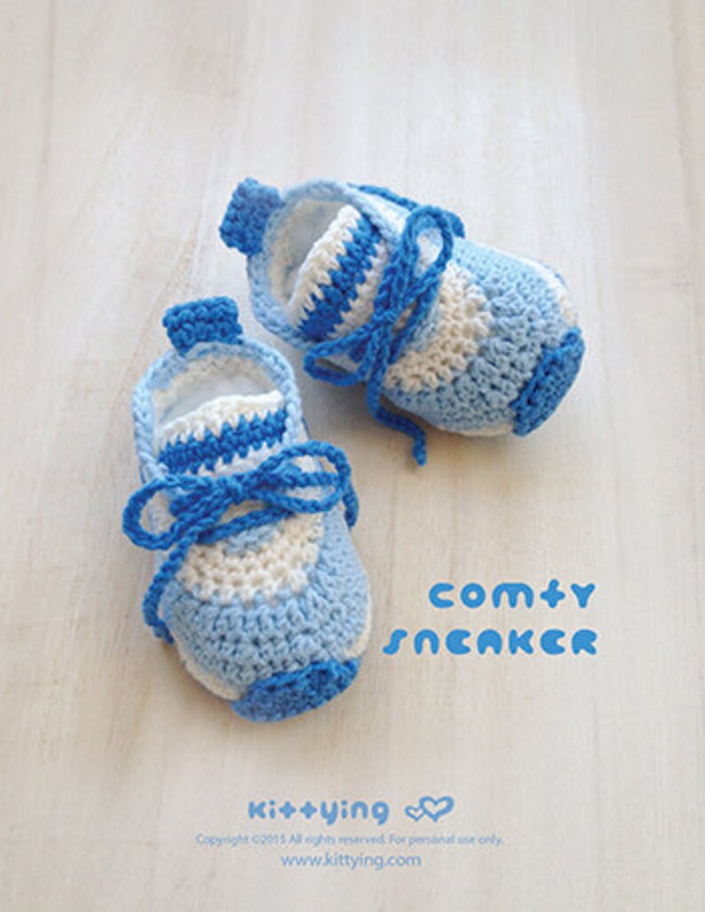May include: A pair of blue and white crocheted baby shoes with laces. The shoes are on a white wooden surface. The text "comty SNCAKCR" is written in blue letters below the shoes.