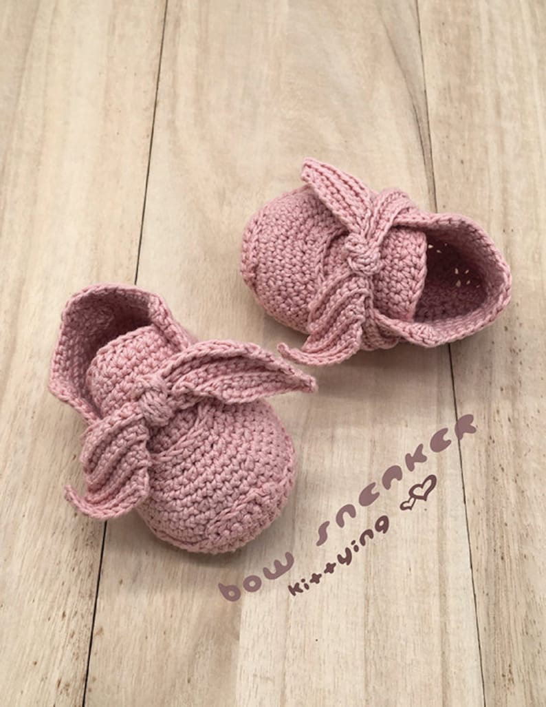May include: A pair of pink crocheted baby shoes with a bow detail. The shoes are on a wooden surface and the text "bow sneaker kit+y.na" is visible in the lower right corner of the image.
