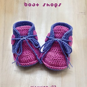 May include: A pair of crocheted baby booties in pink and purple with a blue lace. The booties have a heart-shaped design on the side. The text "bost shops" and "Ki++ying" is visible in the image.