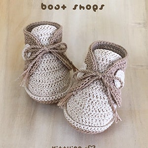 May include: A pair of crocheted baby booties in beige and white with a brown ribbon tie. The booties have a textured knit pattern and are labeled "boot shops" and "kit+ying".