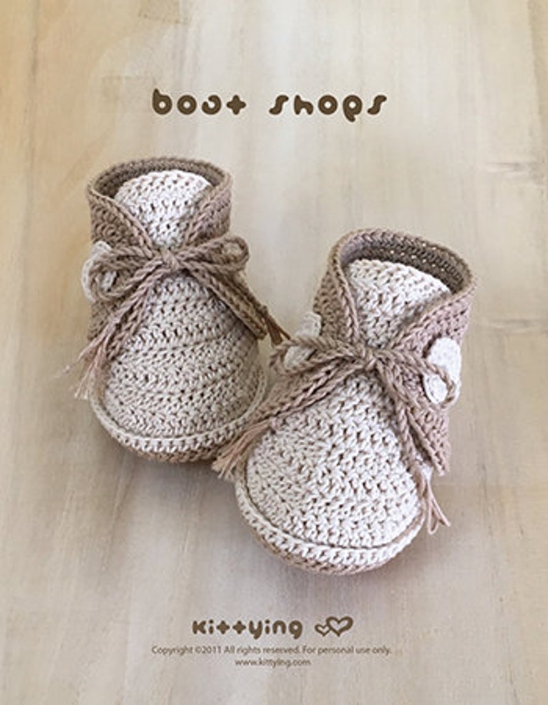 May include: A pair of crocheted baby booties in beige and white with a brown ribbon tie. The booties have a textured knit pattern and are labeled "boot shops" and "kit+ying".