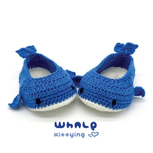 May include: A pair of blue crocheted whale baby shoes with white accents. The shoes have black eyes and a white mouth. The text "WHALE kit+ying" is printed below the shoes.