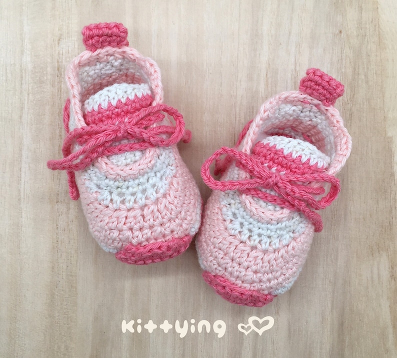 May include: A pair of pink and white crocheted baby booties with a pink ribbon tied around the ankle. The booties have a white crocheted design on the front.