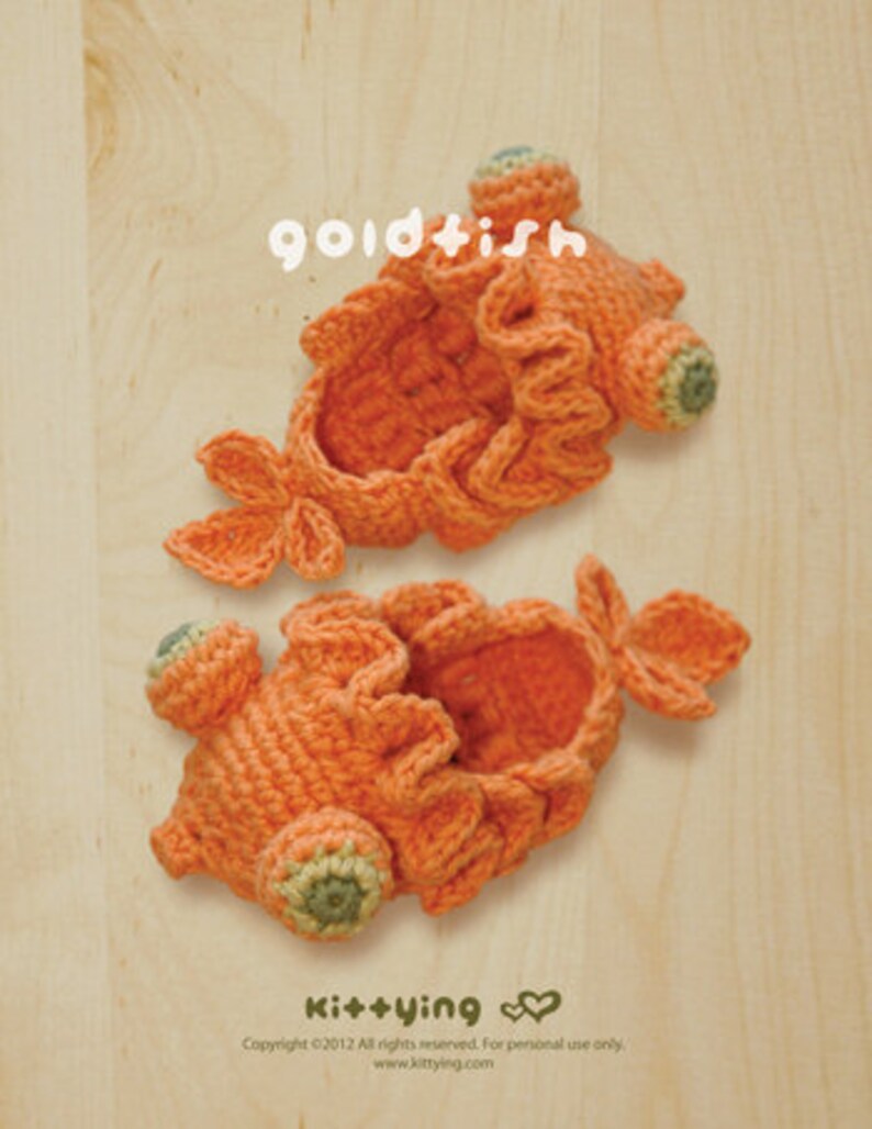 May include: Two handmade orange crochet goldfish, with green and orange accents, are displayed on a light wooden surface. The word "goldfish" is written in white script above the fish. The bottom right corner has the text "kittying" and copyright information.