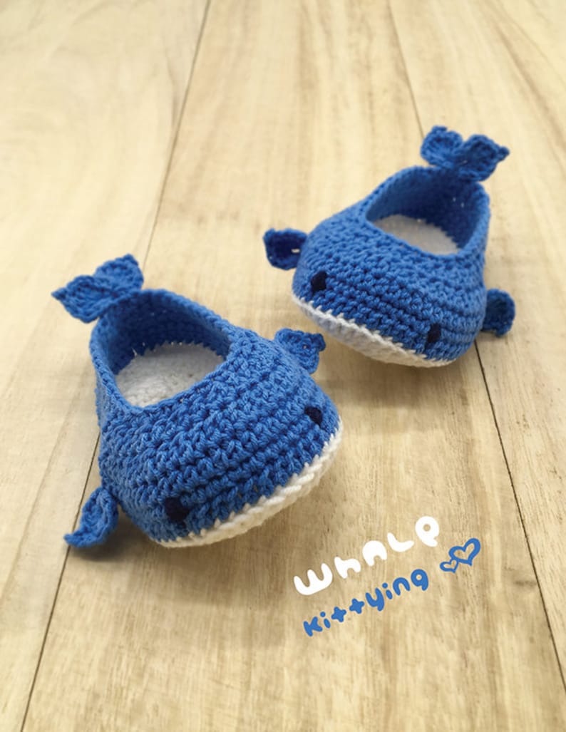 Puede incluir: Un par de zapatos de beb&eacute; de ballena de crochet azul con detalles blancos. Los zapatos tienen un dise&ntilde;o simple con una cola de ballena y una boca blanca. Los zapatos est&aacute;n sobre una superficie de madera con el texto "WHALE Kit+Ying &hearts;" escrito debajo.