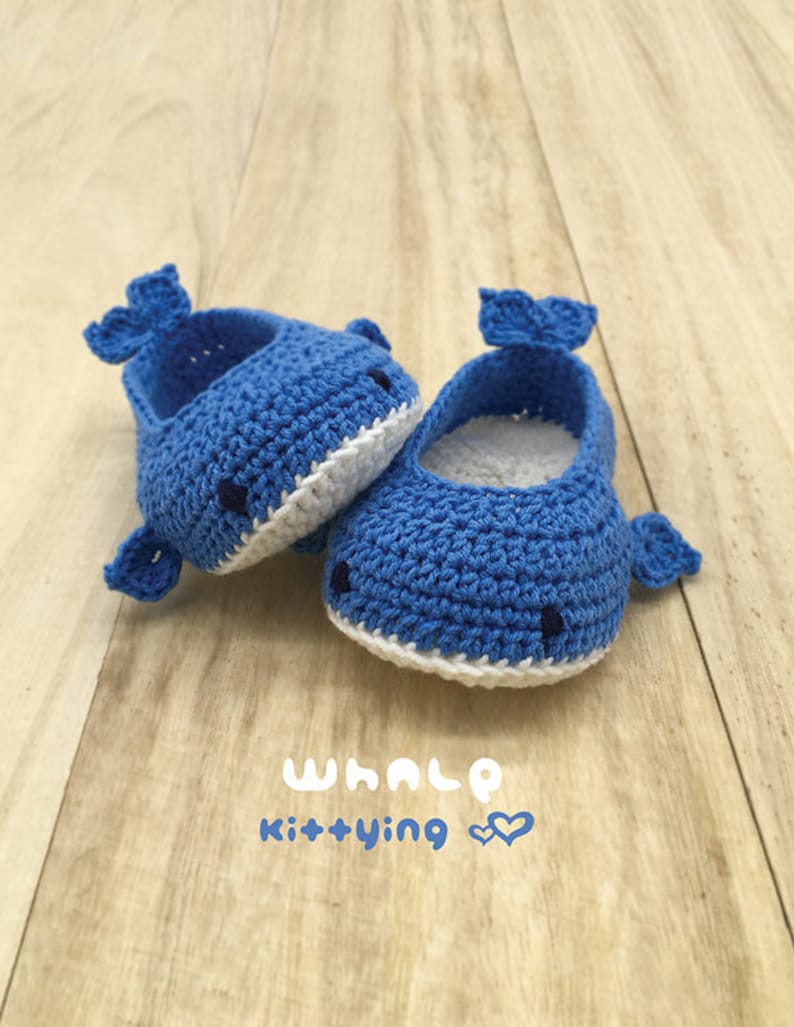 Puede incluir: Un par de zapatos de beb&eacute; de ballena de crochet azul con detalles blancos. Los zapatos est&aacute;n sobre una superficie de madera. El texto "WHALE kit+ying" est&aacute; escrito debajo de los zapatos.