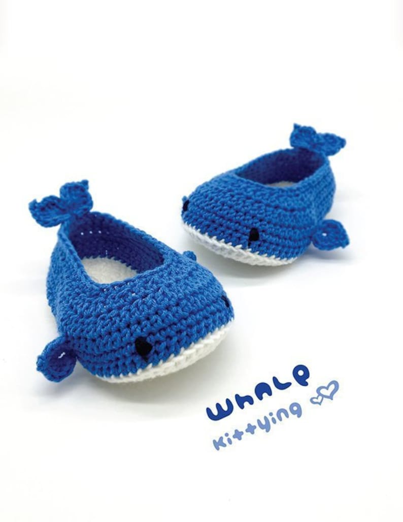 Puede incluir: Un par de zapatos de beb&eacute; de ballena tejidos a crochet en azul con detalles blancos. Los zapatos tienen un dise&ntilde;o simple con una cola y una aleta de ballena en cada zapato. El texto "WHALE Kit+Ying" est&aacute; escrito en la superficie blanca debajo de los zapatos.
