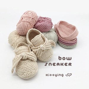 May include: A pair of crocheted baby shoes with a bow detail. The shoes are beige and are surrounded by balls of yarn in various shades of pink and green. The text "bow sneaker kit+ying" is written below the shoes.