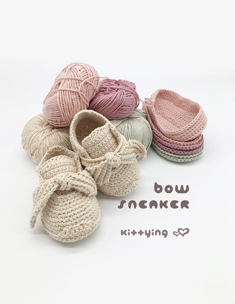 May include: A pair of crocheted baby shoes with a bow detail. The shoes are beige and are surrounded by balls of yarn in various shades of pink and green. The text "bow sneaker kit+ying" is written below the shoes.