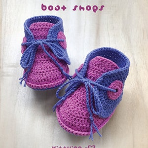 May include: A pair of purple and blue crocheted baby booties with a button detail. The booties are tied with a purple ribbon. The text "bost shops" and "Kit+ying" is visible in the image.