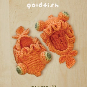 May include: Two orange crocheted goldfish-shaped items with ruffled edges and green accents. The word "goldfish" is written in white script above the items. The items are on a light wood-grain surface.