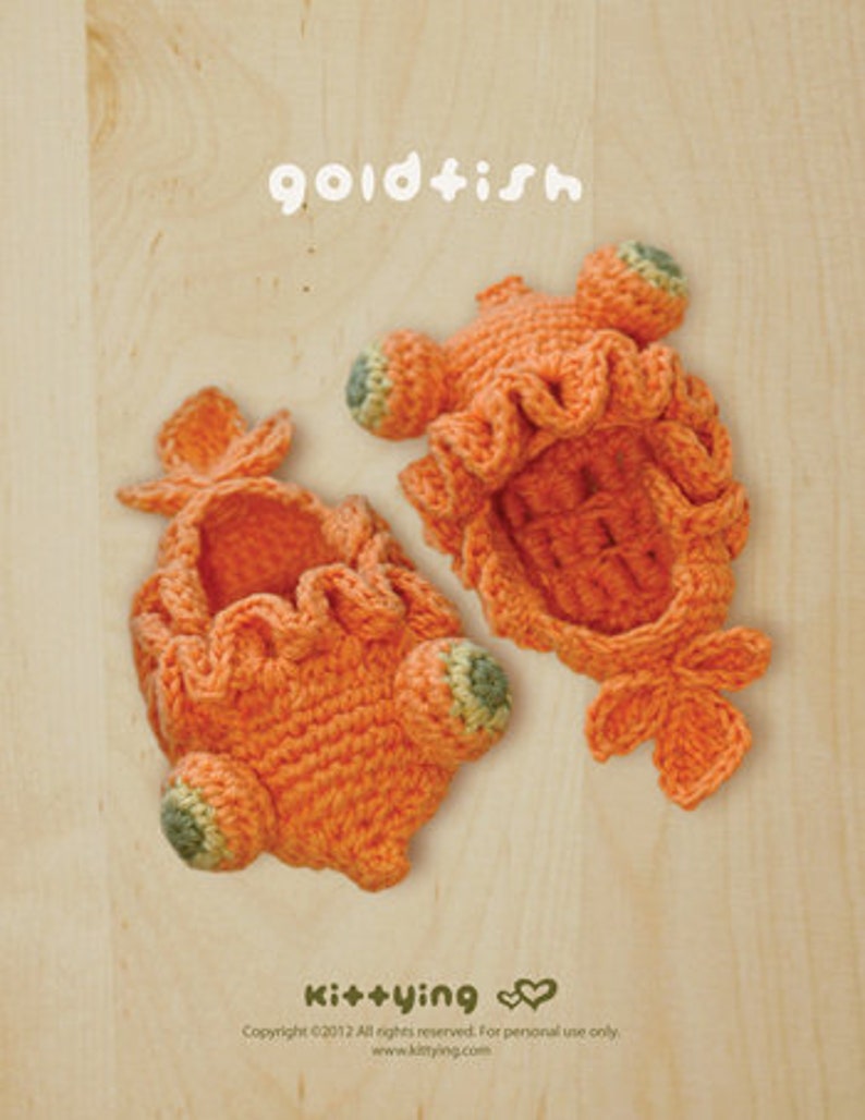 May include: Two orange crocheted goldfish-shaped items with ruffled edges and green accents. The word "goldfish" is written in white script above the items. The items are on a light wood-grain surface.