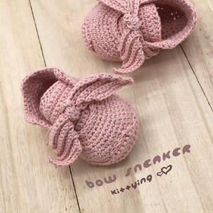 May include: A pair of pink crocheted baby shoes with a bow on the front. The shoes are on a wooden surface and the text "bow sneaker kittying" is written below them.