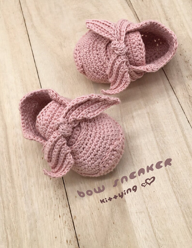 May include: A pair of pink crocheted baby shoes with a bow on the front. The shoes are on a wooden surface and the text "bow sneaker kittying" is written below them.