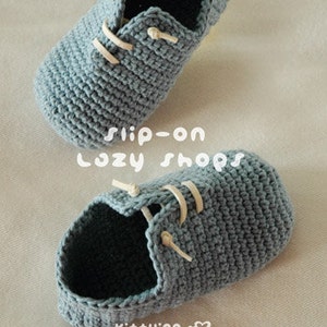 May include: A pair of gray crocheted slip-on shoes with white laces. The shoes are made with a simple stitch pattern and have a rounded toe.