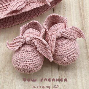 May include: A set of pink crocheted baby shoes with a bow detail. The shoes are on a light wood surface. The text "bow sneaker kit+ying" is visible below the shoes.