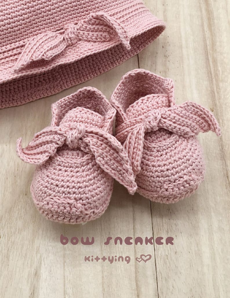 May include: A set of pink crocheted baby shoes with a bow detail. The shoes are on a light wood surface. The text "bow sneaker kit+ying" is visible below the shoes.