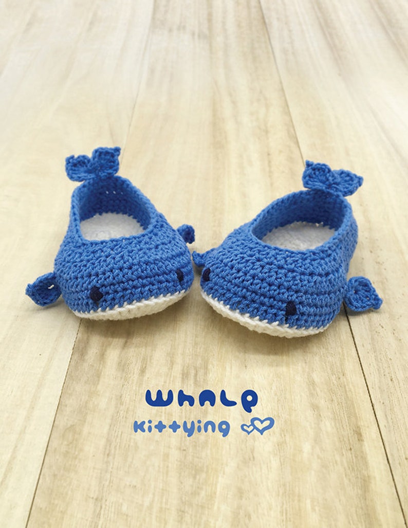 Puede incluir: Un par de zapatos de beb&eacute; de crochet azul con forma de ballenas. Los zapatos tienen detalles blancos y ojos negros. El texto "WHALE Kittying" est&aacute; impreso debajo de los zapatos.