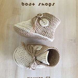 May include: A pair of crocheted baby booties in beige and cream with a button detail. The booties are tied with a beige ribbon and have the text "bost shops" and "Kit+ying" on the image.