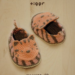 May include: A pair of crocheted baby slippers in the shape of a tiger. The slippers are brown and pink with a tiger face on the front. The slippers are on a wooden surface.