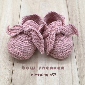 May include: A pair of pink crocheted baby shoes with a bow on the top. The shoes are on a wooden surface. The text "bow sneaker kit+ying" is written below the shoes.
