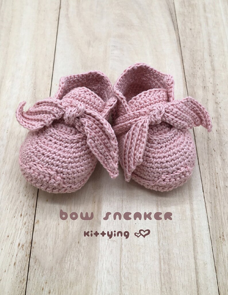 May include: A pair of pink crocheted baby shoes with a bow on the top. The shoes are on a wooden surface. The text "bow sneaker kit+ying" is written below the shoes.