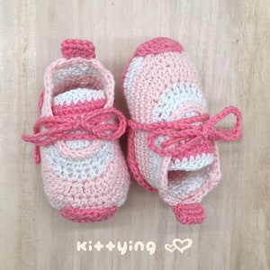 May include: A pair of pink and white crocheted baby shoes with pink laces tied in a bow. The shoes have a textured design and are made with a soft yarn.