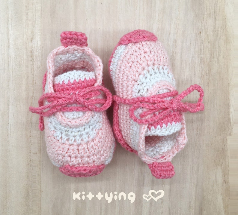 May include: A pair of pink and white crocheted baby shoes with pink laces tied in a bow. The shoes have a textured design and are made with a soft yarn.