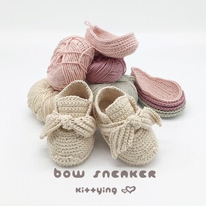 May include: A pair of beige crocheted baby shoes with a bow on the front. The shoes are surrounded by balls of yarn in various shades of pink and beige. The text "bow sneaker kit+ying &hearts;&hearts;" is written below the shoes.