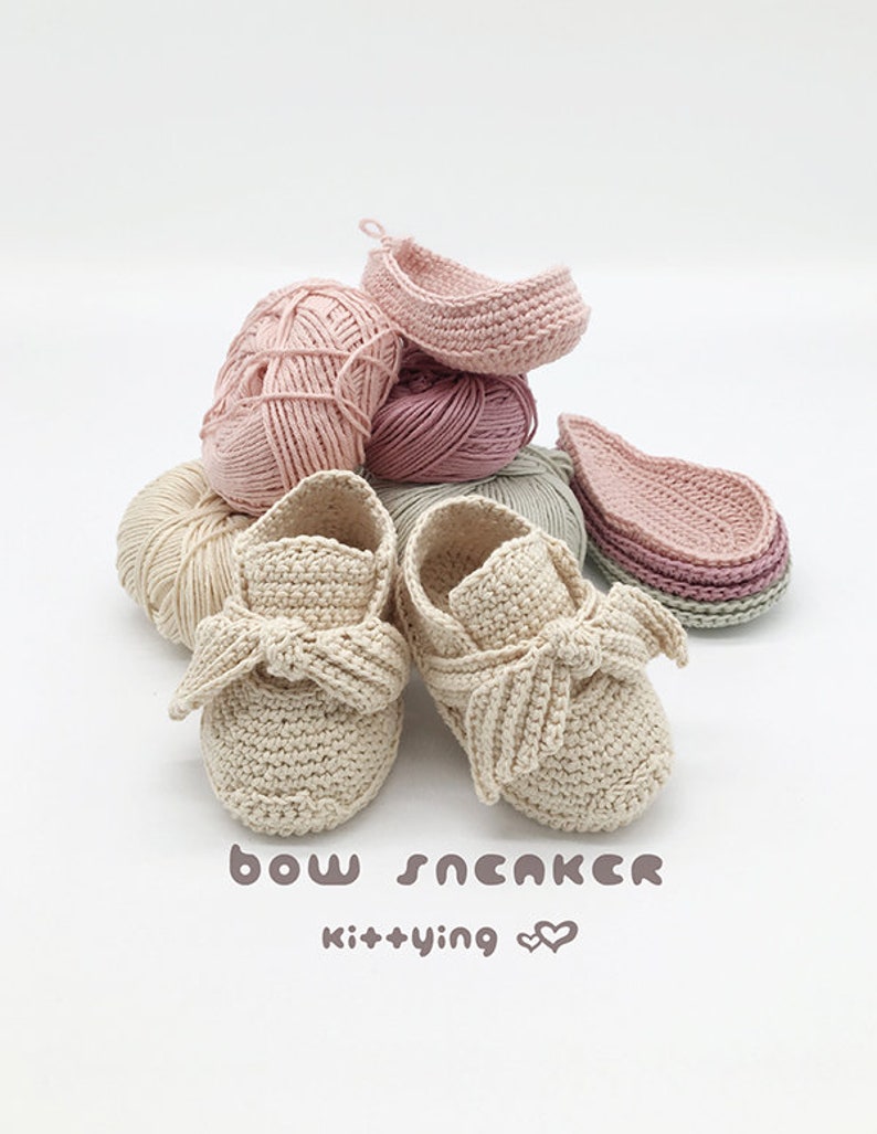 May include: A pair of beige crocheted baby shoes with a bow on the front. The shoes are surrounded by balls of yarn in various shades of pink and beige. The text "bow sneaker kit+ying &hearts;&hearts;" is written below the shoes.
