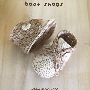 May include: A pair of crocheted baby booties in beige and cream with a button detail and a tassel on each shoe. The booties are laid out on a wooden surface. The text "boat shops" is at the top of the image and the text "Ki++ying" is at the bottom of the image.