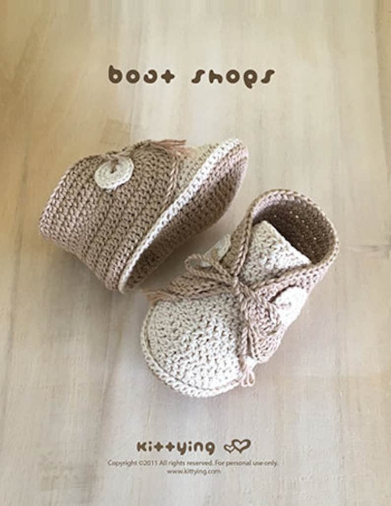 May include: A pair of crocheted baby booties in beige and cream with a button detail and a tassel on each shoe. The booties are laid out on a wooden surface. The text "boat shops" is at the top of the image and the text "Ki++ying" is at the bottom of the image.