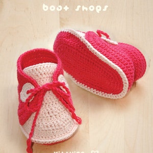 May include: A pair of pink and white crocheted baby booties with a white lace trim. The booties have a white heart on the front and the text "boo+shops" is written above the booties.