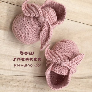May include: A pair of pink crocheted baby booties with a bow detail. The booties are on a wooden surface. The text "bow sneaker kittying" is written on the surface.