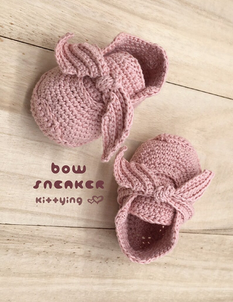 May include: A pair of pink crocheted baby booties with a bow detail. The booties are on a wooden surface. The text "bow sneaker kittying" is written on the surface.