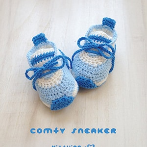 May include: A pair of blue and white crocheted baby sneakers with blue laces. The sneakers are on a white wooden surface. The text "comty SNEAKER" and "Ki++ying" is visible below the sneakers.