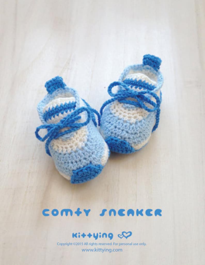 May include: A pair of blue and white crocheted baby sneakers with blue laces. The sneakers are on a white wooden surface. The text "comty SNEAKER" and "Ki++ying" is visible below the sneakers.