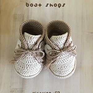 May include: A pair of beige and brown crocheted baby booties with a bow detail. The booties are labeled "boot shops" and "Kit+ying".