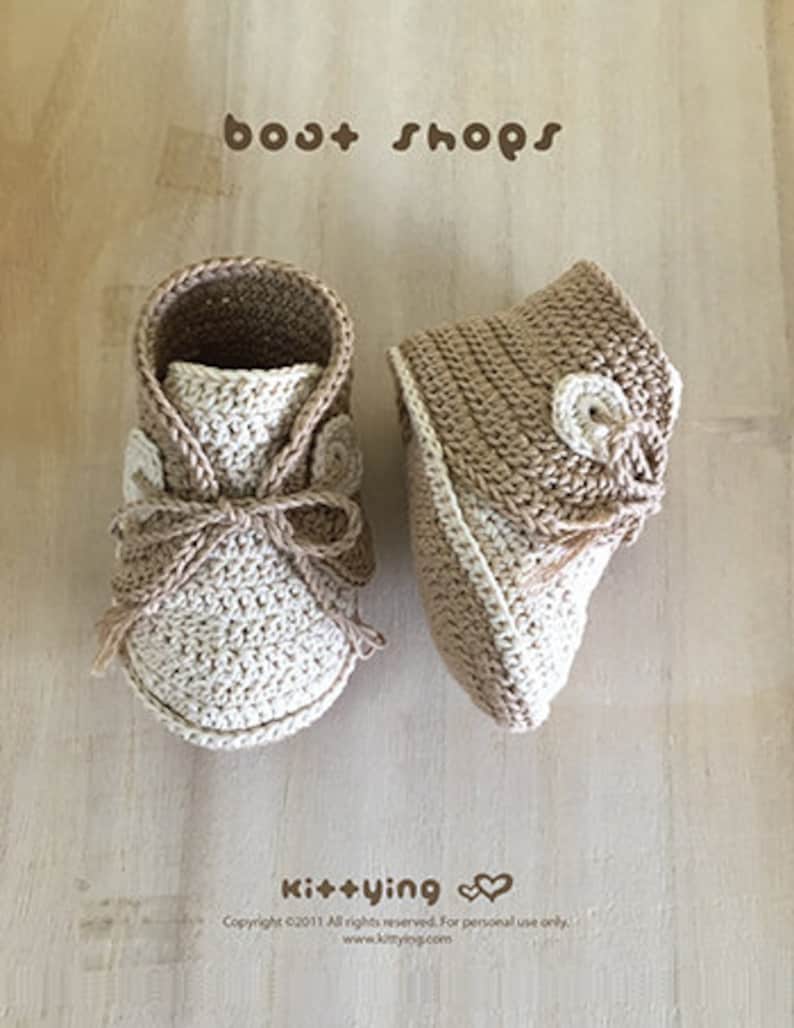 May include: A pair of crocheted baby booties in beige and cream with a button detail. The booties are on a wooden surface. The text "boat shops" is at the top of the image and the text "Ki++ying" is at the bottom of the image.