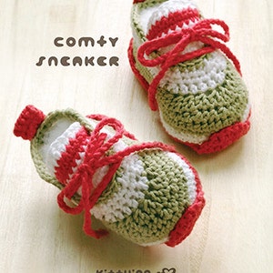 Könnte beinhalten: Ein Paar gehäkelte Babyschuhe in Grün, Weiß und Rot. Die Schuhe haben Schnürsenkel und eine abgerundete Spitze. Der Text "comty SNCAKCR" und "Ki++ying" ist im Bild sichtbar.