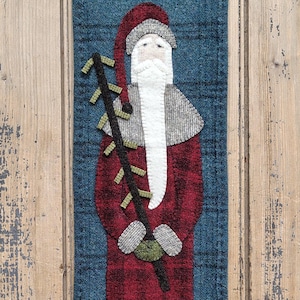 May include: A festive Christmas wall hanging featuring a felt Santa Claus design. Santa is depicted in a red plaid coat, a white beard, and a red hat. He holds a black staff with green accents against a blue background.