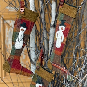 May include: Three handmade felt Christmas stockings with a snowman design in shades of red, green, brown, and gold hanging on a birch branch.