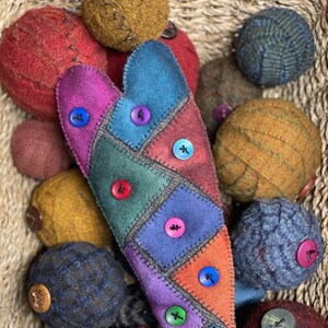 Op de afbeelding: Een hartvormige patchwork-versiering met kleurrijke vierkanten van stof en knopen. De versiering is omringd door een verscheidenheid aan wollen ballen in verschillende kleuren en texturen.