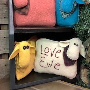 Peut inclure: Quatre coussins en forme de mouton en feutre, deux sont roses, un est bleu, un est jaune et un est blanc avec le texte "Love ewe" brodé dessus.