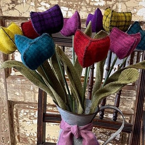 May include: A bouquet of felt tulips in various colours, including purple, red, yellow, teal, and green, arranged in a galvanised metal pitcher with a purple ribbon. The pitcher is sitting on a colourful braided rug.